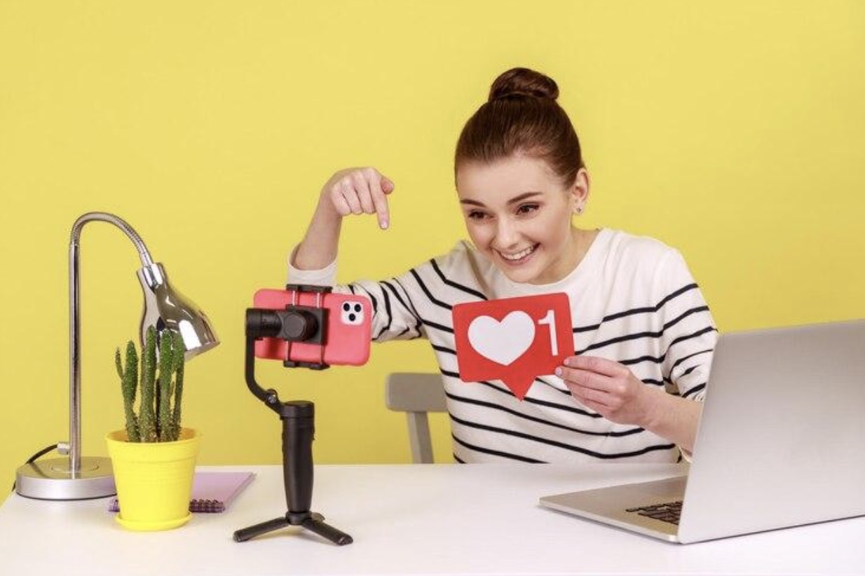 Woman blogger recording video for followers holding like sign asking to rate her posts and subscribe